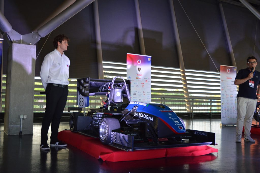 Blue and black race car on a red pedestal, two men nearby at an indoor auto show with banners in the background.